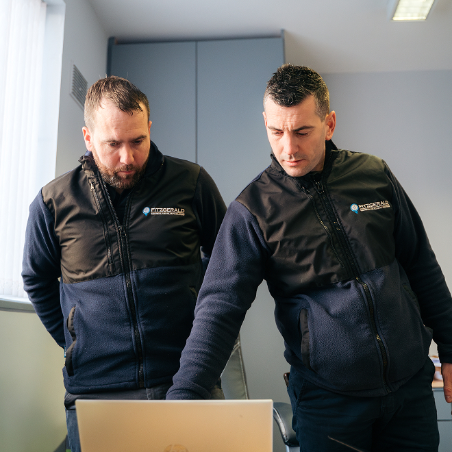 Two men, standing, in Fitzgerald Energy Solutions uniform examining a laptop display in an office setting.